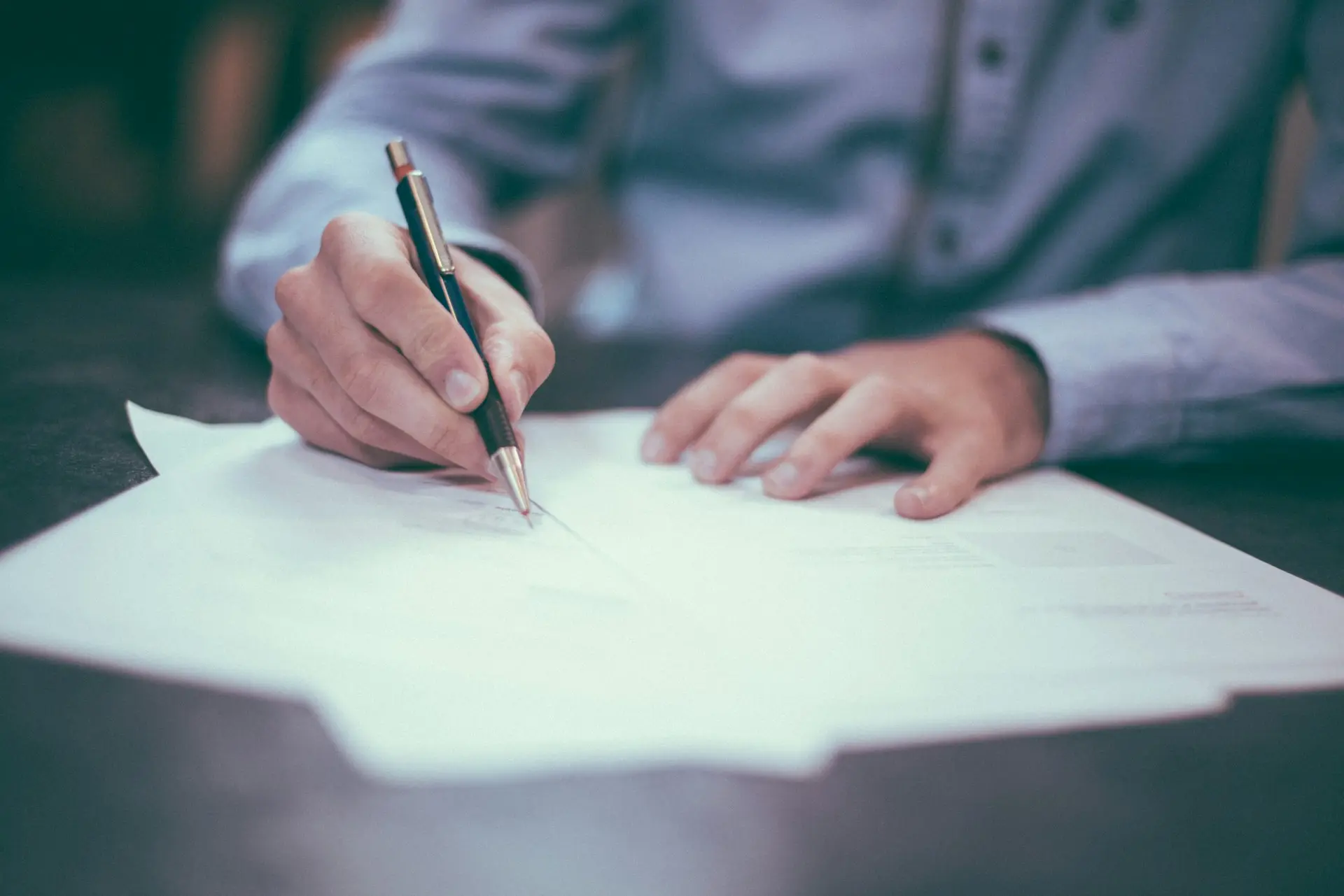 Signature ou Révision de Documents dans un Cadre Professionnel Gros plan d’un professionnel signant ou examinant des documents avec un stylo sur un bureau, vêtu d’une chemise bleue.