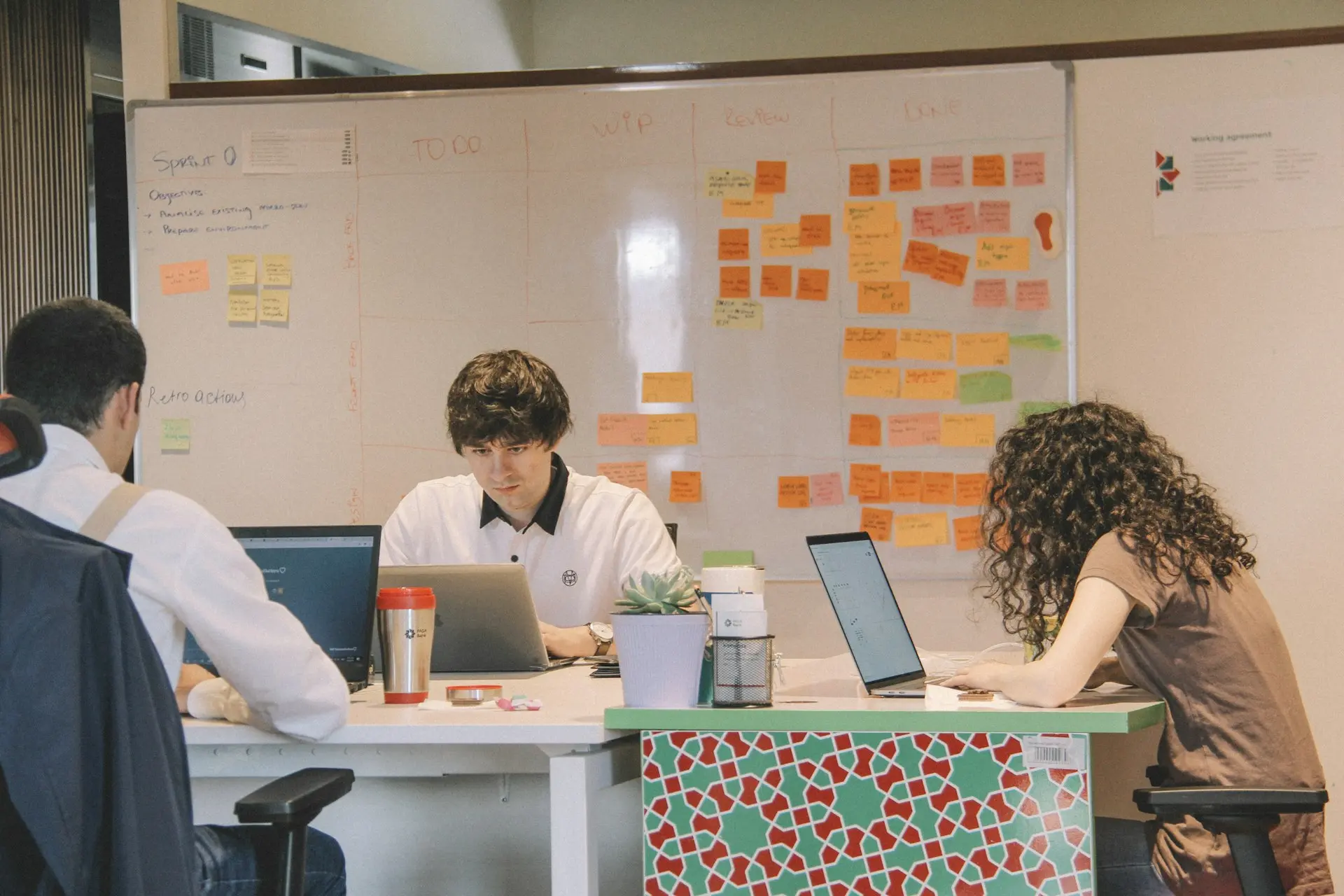 Équipe Collaborative Travaillant avec un Tableau Agile Trois professionnels travaillant à un bureau dans un bureau collaboratif, avec un tableau blanc recouvert de notes autocollantes colorées pour la gestion des tâches.