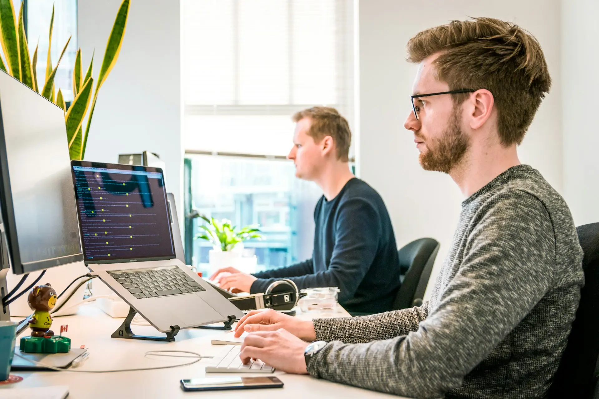 Geconcentreerde Professionals aan het Werk in een Modern Kantoor Twee geconcentreerde professionals aan het werk op computers in een modern, licht kantoor met grote ramen en kamerplanten.
